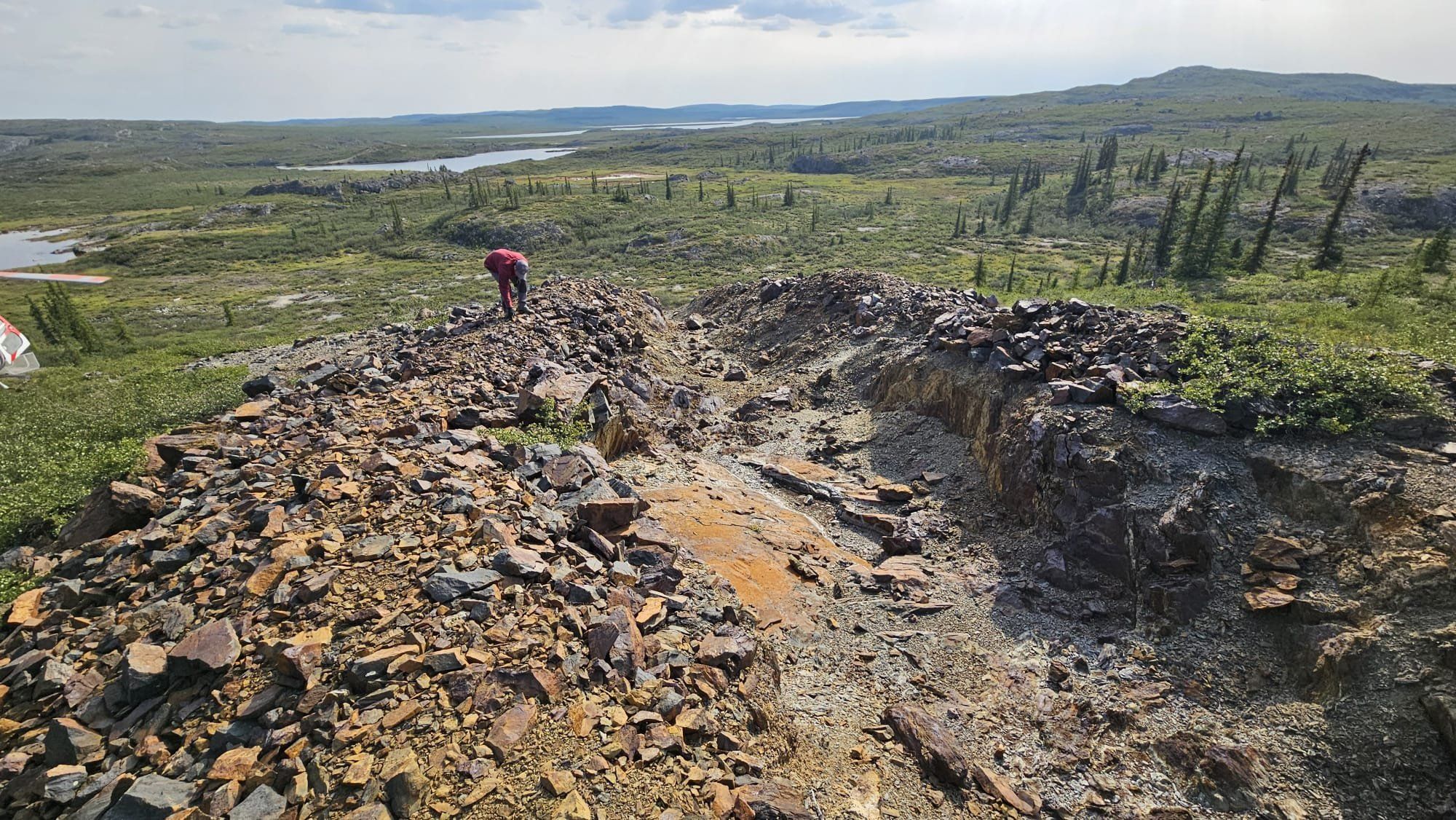 A geologist sampling a rocky outcrop on the Olympus property, with lakes and spruce forest behind
